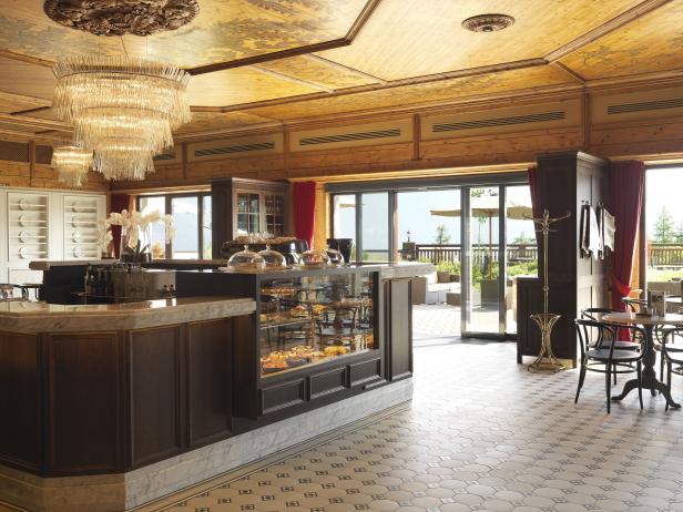 Innenansicht eines Cafés mit Holztäfelung, einer Theke mit Kuchenauslage und einem Kronleuchter.