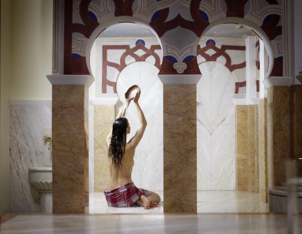 Eine Frau sitzt kniend in einem Hamam und gießt Wasser über ihren Kopf.
