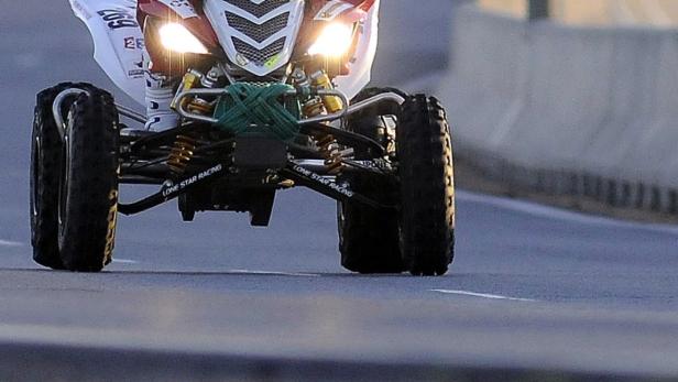 Ein Quad mit eingeschalteten Scheinwerfern fährt auf einer asphaltierten Straße.