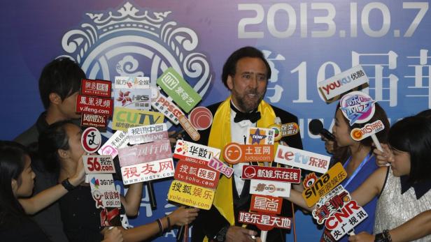 Nicolas Cage umringt von Reportern mit Mikrofonen und Logos verschiedener Medienunternehmen.
