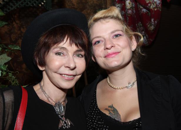 Zwei Frauen posieren nebeneinander, die eine mit Hut und auffälliger Halskette, die andere mit einem Tattoo auf der Brust.