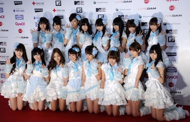 Eine Gruppe japanischer Mädchen in blau-weißen Kostümen kniet vor einer Wand mit Sponsorenlogos.