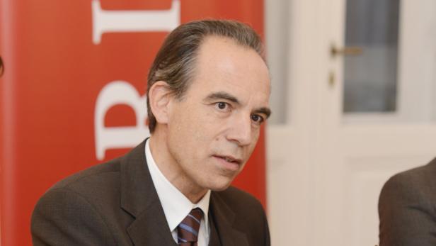 Ein Mann in einem dunklen Anzug und Krawatte vor einem roten Hintergrund.