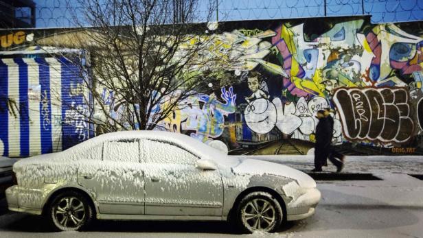 Ein verschneites Auto steht vor einer mit Graffiti bemalten Wand, während eine Person vorbeigeht.