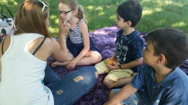 Eine Frau schminkt ein Mädchen mit Noten im Gesicht, während zwei Jungen zusehen.