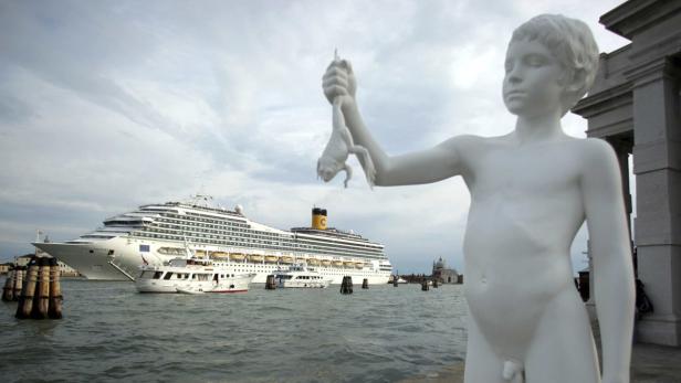 Eine Statue eines Jungen, der einen Frosch in Venedig hält, mit einem Kreuzfahrtschiff im Hintergrund.