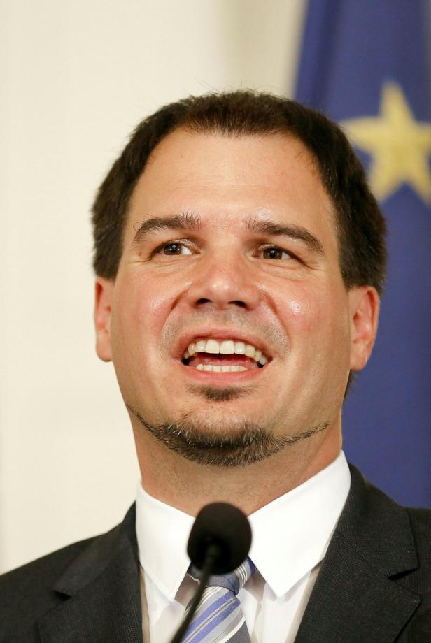 Ein Mann mit Anzug und Krawatte spricht vor einer Europaflagge.