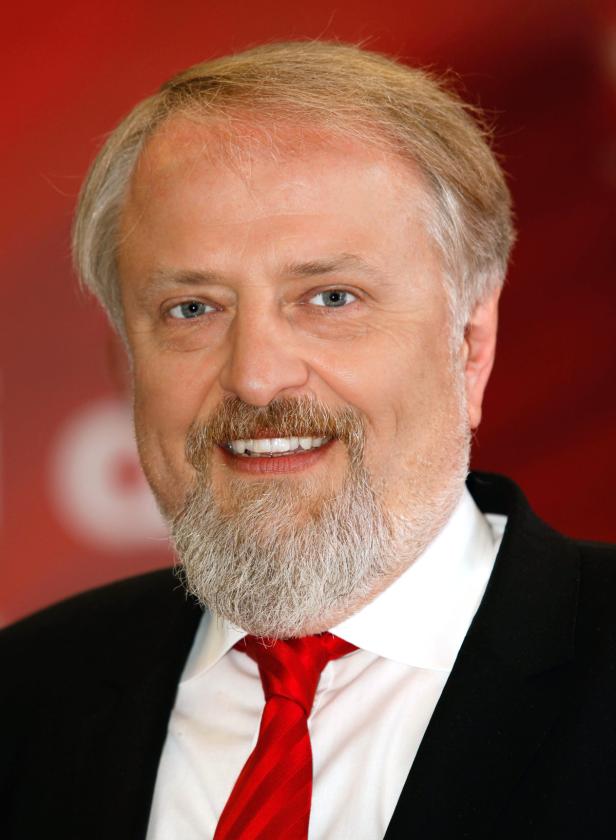 Porträt eines lächelnden Mannes mit Bart und roter Krawatte vor rotem Hintergrund.