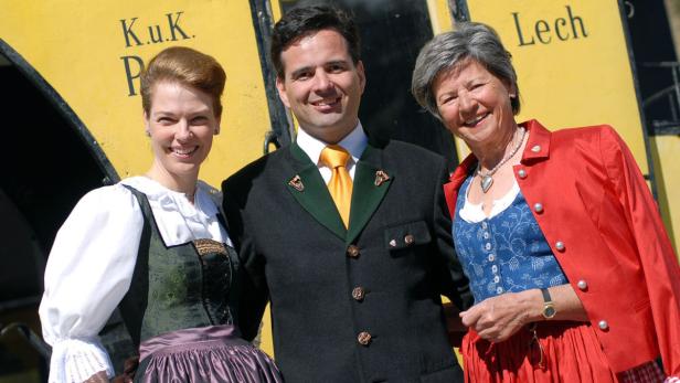 Drei Personen in Tracht posieren vor einem gelben Zug der K.u.K. Post in Lech.