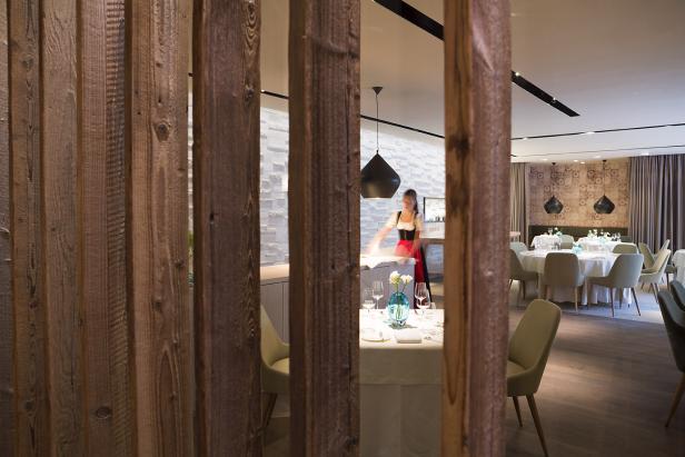 Blick durch eine hölzerne Trennwand auf ein Restaurant mit einer Kellnerin in Tracht.