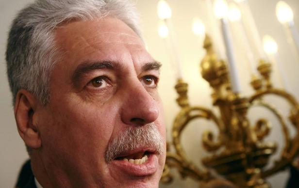 Nahaufnahme eines Mannes mit grauem Haar und Schnurrbart vor einem goldenen Kerzenständer.