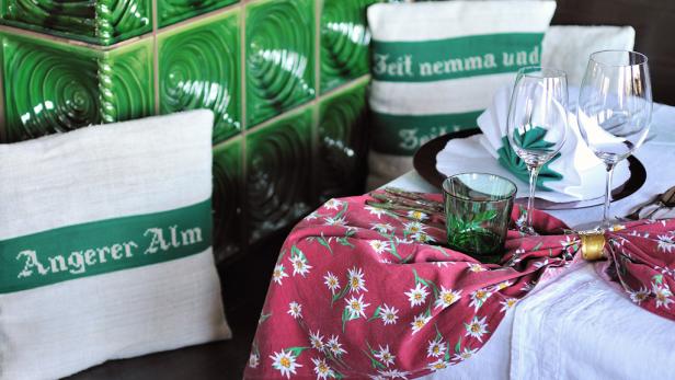 Ein gedeckter Tisch mit Gläsern, Servietten und Kissen mit der Aufschrift „Angerer Alm“.