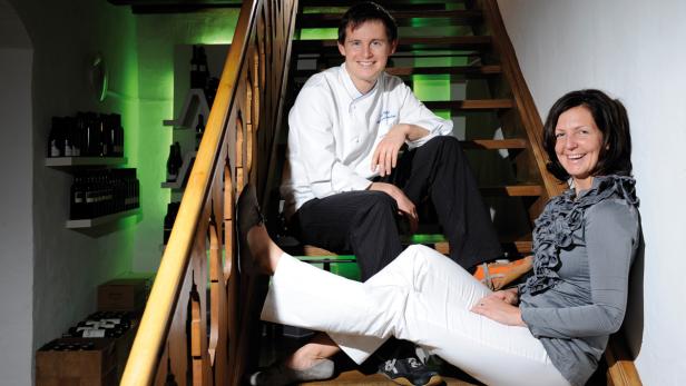 Ein Koch und eine Frau sitzen lächelnd auf einer Holztreppe vor einem Weinregal.