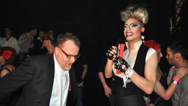 Eine Dragqueen mit extravaganten Make-up und ein Mann im Anzug auf einer Party.