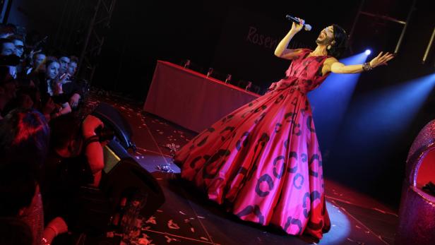 Conchita Wurst steht in einem roten Kleid auf einer Bühne und singt.