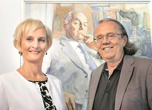 Zwei Personen stehen vor einem Porträtgemälde eines Mannes.