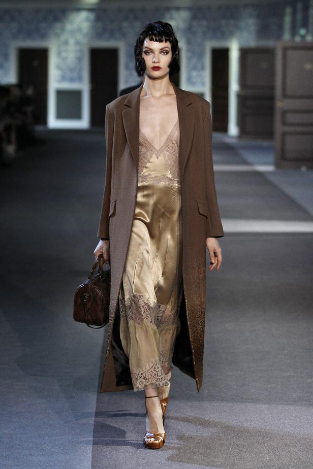 Eine Frau in einem braunen Mantel und einem goldenen Kleid mit Spitze läuft über den Laufsteg.