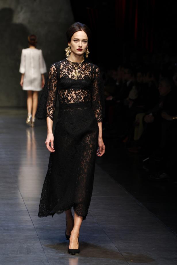 Eine Frau in einem schwarzen Spitzenkleid auf einem Laufsteg mit goldenem Schmuck.