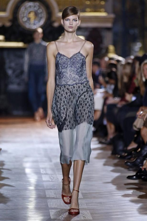 Ein Model präsentiert ein blaues Spitzenkleid und rote Sandalen auf dem Laufsteg.