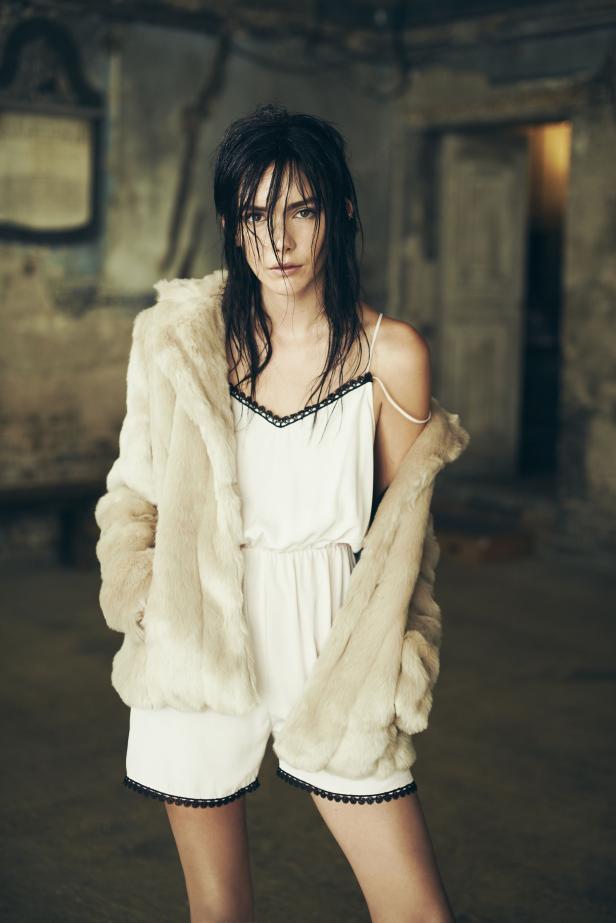 Eine Frau mit nassen Haaren trägt einen weißen Jumpsuit und eine helle Pelzjacke.