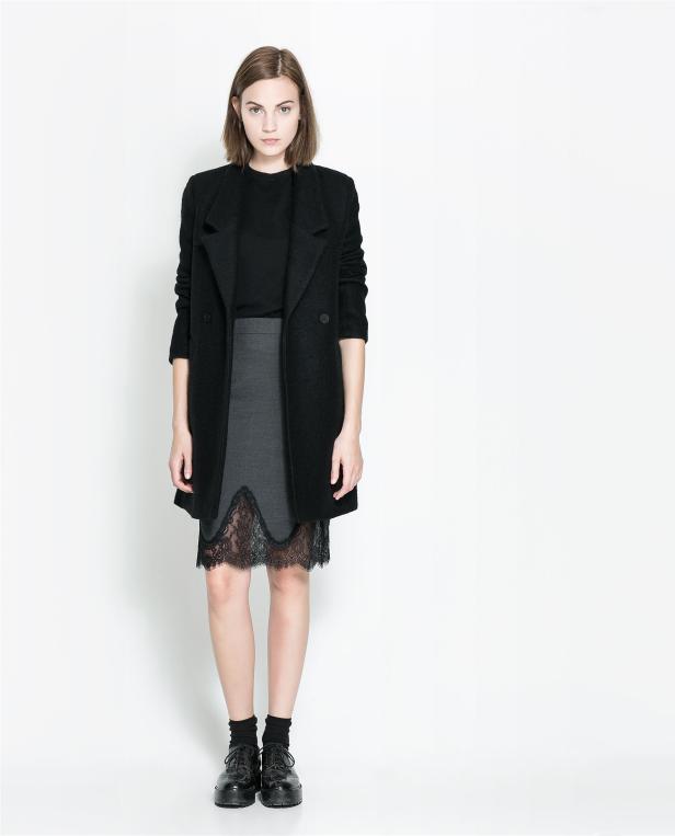 Eine Frau mit kurzem Haar trägt einen schwarzen Mantel und einen grauen Rock mit Spitze.