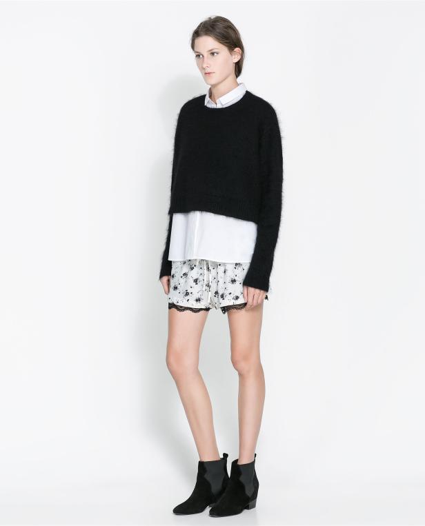 Eine Frau trägt einen schwarzen Pullover, ein weißes Hemd, Shorts mit Blumenmuster und Stiefeletten.