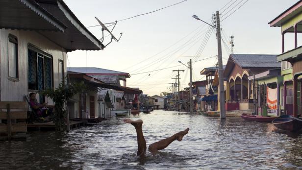 Überflutete Straße in einer Stadt mit Stelzenhäusern, im Vordergrund ragen Beine aus dem Wasser.