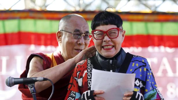 Der Dalai Lama legt seine Hand auf die Schulter einer lächelnden Frau mit roter Brille.