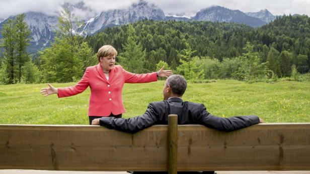 Angela Merkel gestikuliert vor Barack Obama, der auf einer Bank vor einer Bergkulisse sitzt.
