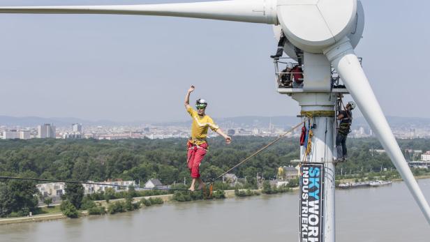 Ein Mann balanciert barfuß auf einem Seil an einem Windrad vor der Skyline von Wien.
