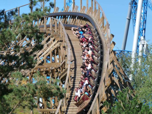 Eine Holzachterbahn ist mit vielen Menschen besetzt und fährt eine steile Kurve hinauf.
