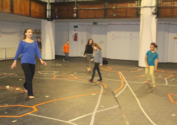Eine Frau leitet eine Gruppe von Kindern barfuß in einem Raum mit Markierungen auf dem Boden an.