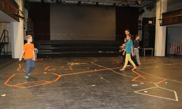 Kinder laufen barfuß auf einer Bühne mit aufgeklebten Markierungen.