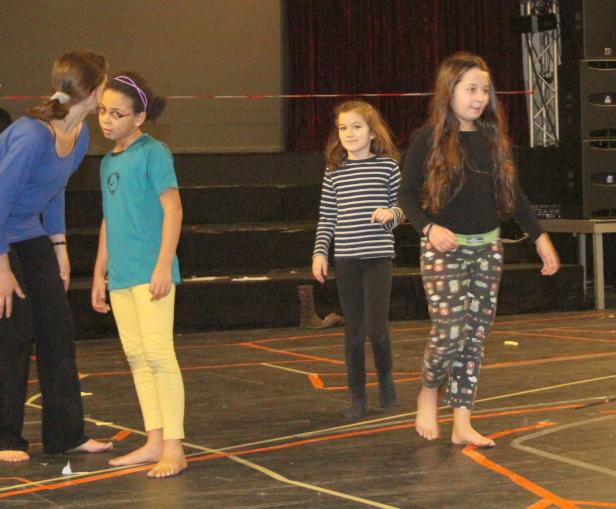 Eine Gruppe barfüßiger Mädchen steht auf einer Bühne mit farbigen Markierungen.