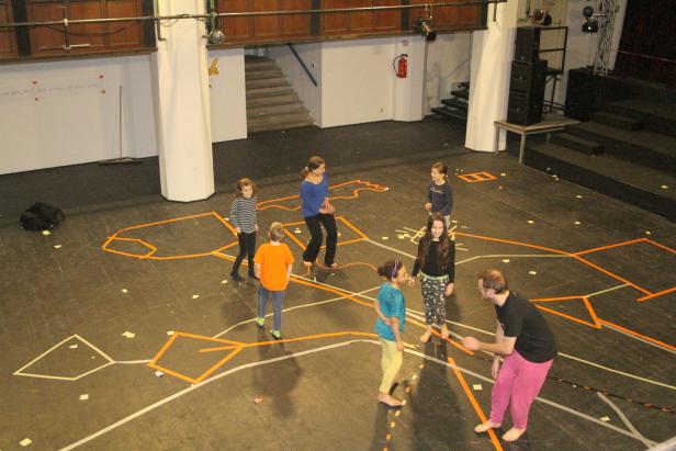 Eine Gruppe von Kindern und ein Erwachsener stehen auf einem Bühnenboden mit aufgeklebten Linien.