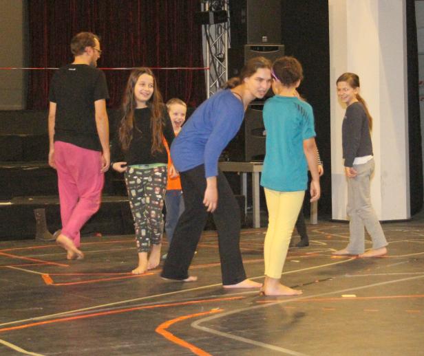 Eine Gruppe von Kindern und ein Erwachsener stehen barfuß auf einer Bühne mit Markierungen.