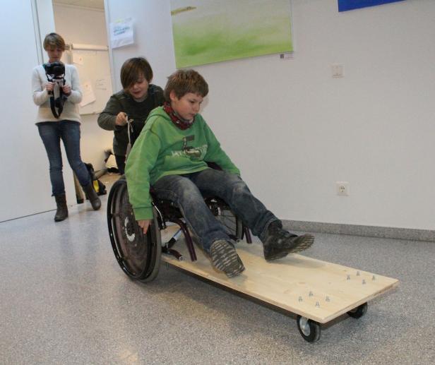 Ein Junge sitzt in einem Rollstuhl auf einem hölzernen Rollbrett, während ein anderer ihn schiebt.