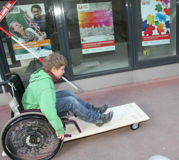 Ein Junge im Rollstuhl sitzt auf einem Holzbrett mit Rollen vor VHS-Plakaten.
