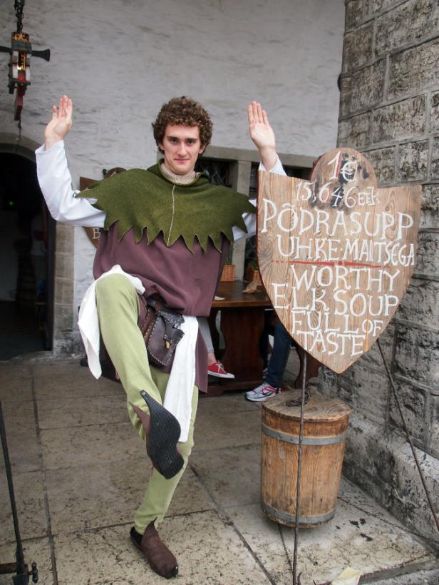 Ein Mann in mittelalterlicher Kleidung posiert neben einem Schild für Elchsuppe.