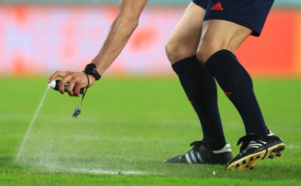 Ein Schiedsrichter sprüht Markierungsspray auf das Gras eines Fußballfeldes.