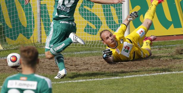 Ein Fußballtorwart hechtet nach dem Ball, während ein Spieler sich nähert.