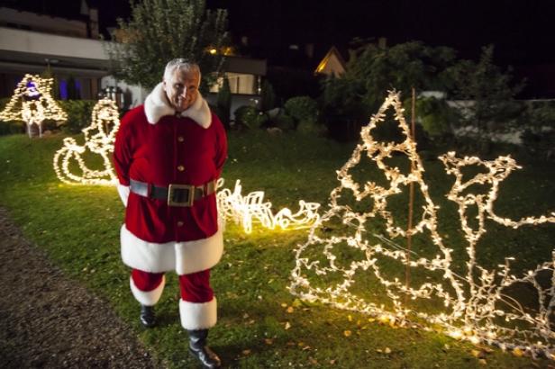 (c: sky österreich) Ein Mann im Weihnachtsmannkostüm steht vor einem Haus mit Weihnachtsbeleuchtung.