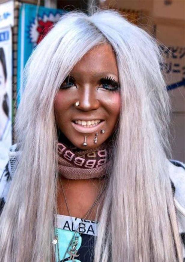 Eine junge Frau mit gebräunter Haut, weißblondem Haar und Piercings im Gesicht lächelt.