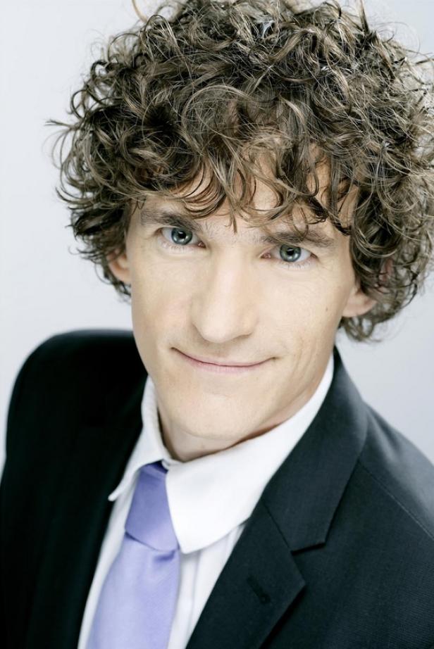 Ein Mann mit lockigem Haar und Anzug lächelt in die Kamera.