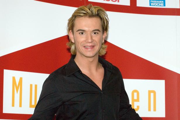 Ein lächelnder Mann mit blonden Haaren und einem schwarzen Hemd vor einem roten und weißen Hintergrund.