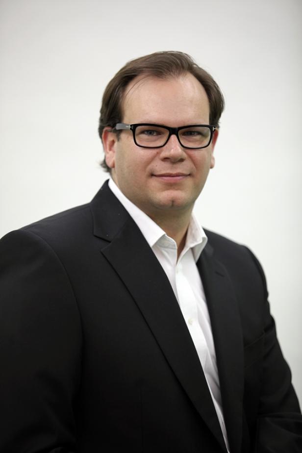 Ein Mann mit Brille und dunklem Anzug vor einem hellen Hintergrund.