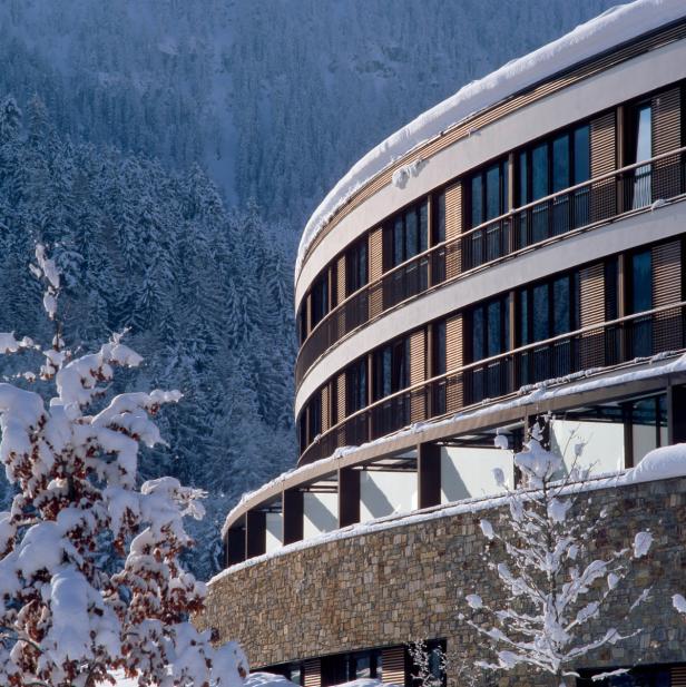 Ein modernes, schneebedecktes Gebäude vor einem bewaldeten Berghang.