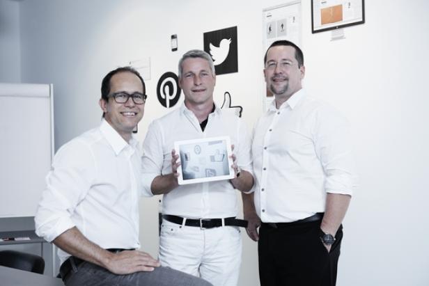 Drei Männer stehen vor einer Wand mit Social-Media-Logos, einer hält ein Tablet.