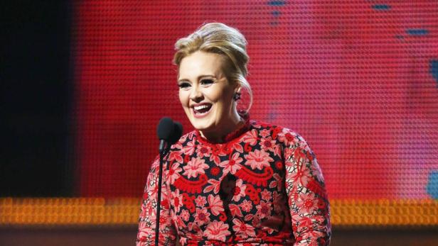 Adele lacht an einem Mikrofon in einem roten, blumengemusterten Kleid.