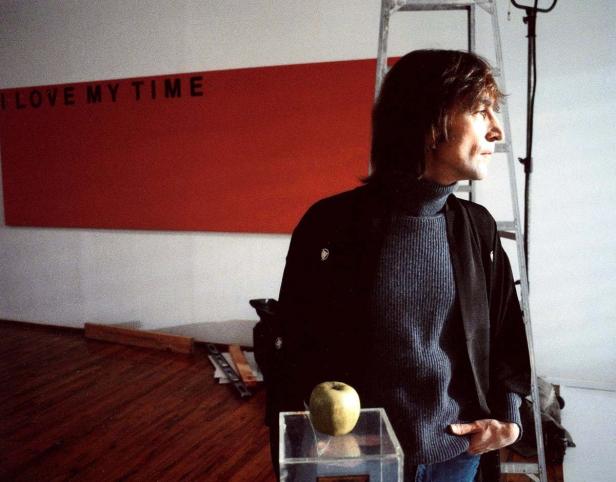 John Lennon steht vor einem roten Bild mit der Aufschrift „I LOVE MY TIME“.